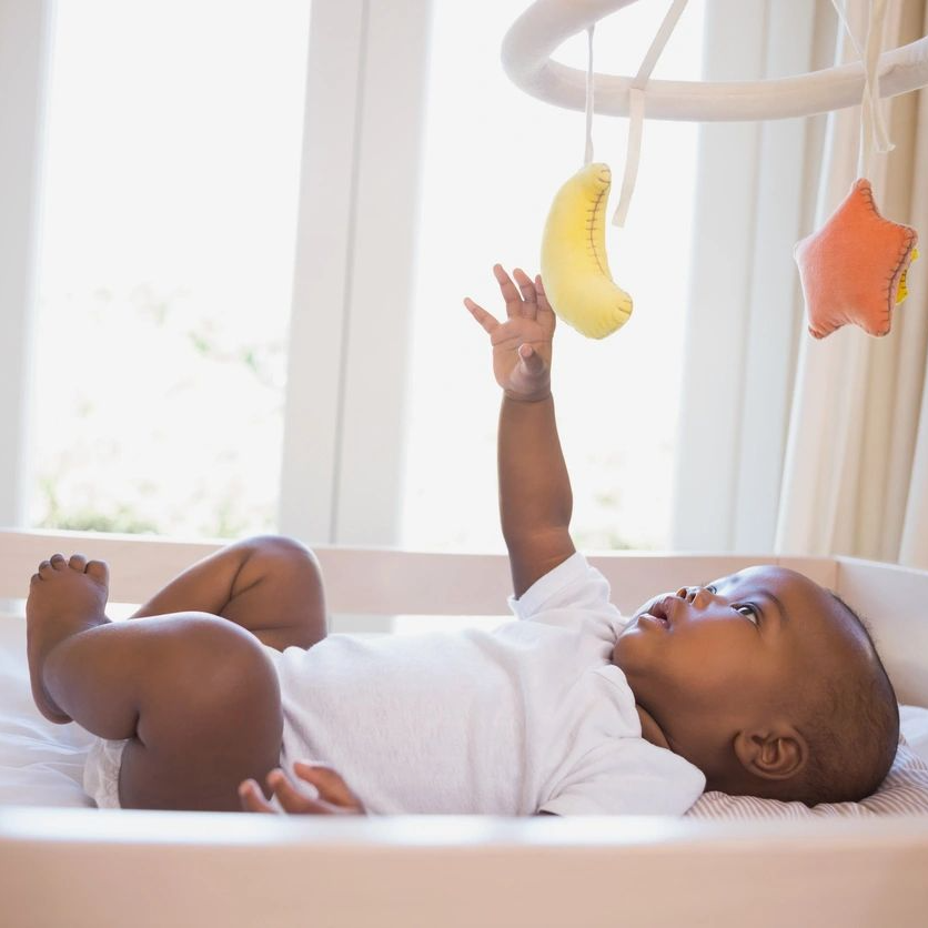 A baby laying in his crib with a banana hanging from the ceiling.