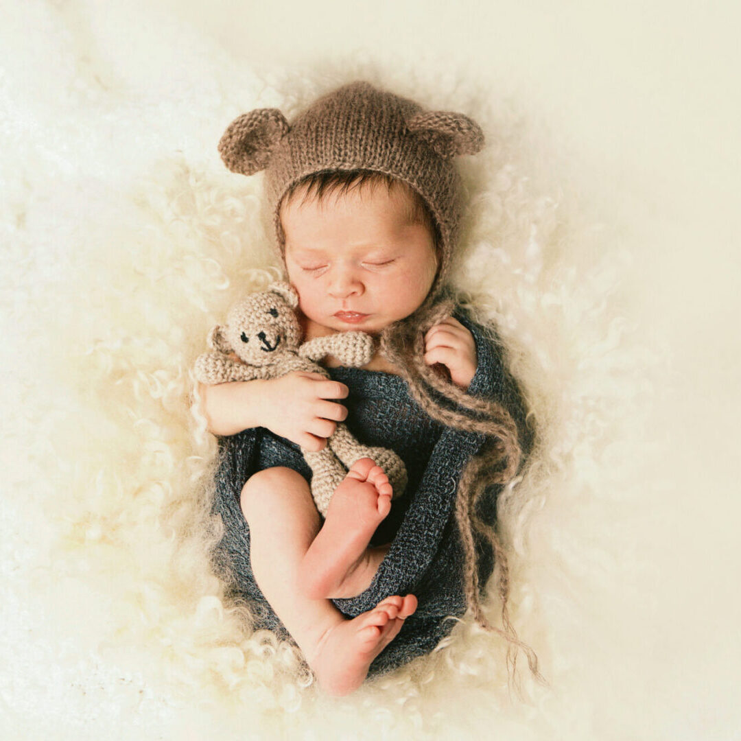 A newborn baby wearing a bonnet with bear ears, snuggling a teddy bear.